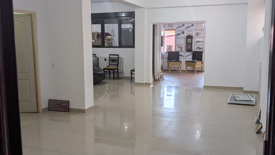 Wide, mostly empty interior room with white walls, a glossy tiled floor, and exposed hanging light bulbs. Open doorways and a supporting column lead to a back area with windows, a small seating setup, and patterned wall panels.
