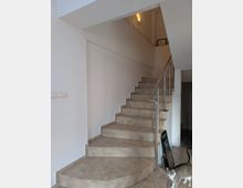 Indoor stairwell with beige tiled steps leading to an upper landing, bordered by white walls and a stainless-steel handrail. A wall sconce light illuminates the landing, with a doorway opening off to the right.