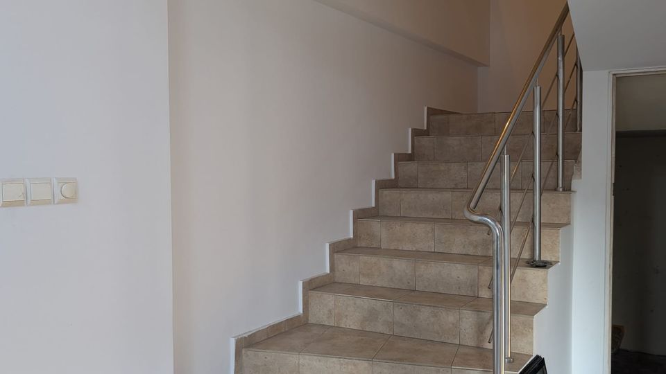 Indoor stairwell with beige tiled steps leading to an upper landing, bordered by white walls and a stainless-steel handrail. A wall sconce light illuminates the landing, with a doorway opening off to the right.