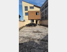 Modern multi-story residential building with a slanted roof and beige facade, featuring a large brown rectangular section and several windows. In front is a paved courtyard driveway with mosaic stone tiles, bordered by walls and an adjacent apartment block.