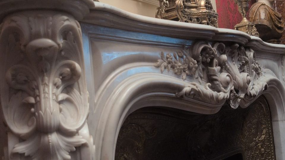 Ornate interior fireplace with a carved white stone mantel featuring floral reliefs, topped with antique metal candelabras and a decorative clock. The firebox has dark, patterned metal panels, and the surrounding walls include red wallpaper and a framed oval artwork near the ceiling.