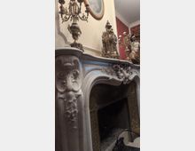 Ornate interior fireplace with a carved white stone mantel featuring floral reliefs, topped with antique metal candelabras and a decorative clock. The firebox has dark, patterned metal panels, and the surrounding walls include red wallpaper and a framed oval artwork near the ceiling.