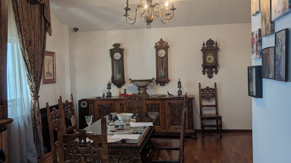 Traditional dining room with a long carved wooden table and matching chairs on a polished wood floor, lit by a hanging chandelier. The far wall has ornate wooden cabinets and several antique wall clocks, with heavy drapes over a window and framed artwork on the side wall.