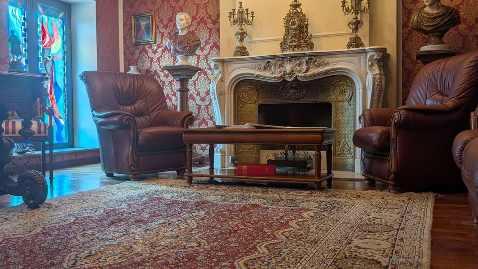 Elegant sitting room with red damask wallpaper, a carved stone fireplace topped with ornate clocks and candelabras, and a central chandelier. Leather armchairs and a coffee table sit on a large patterned Persian-style rug, with a stained-glass window and decorative busts along the walls.